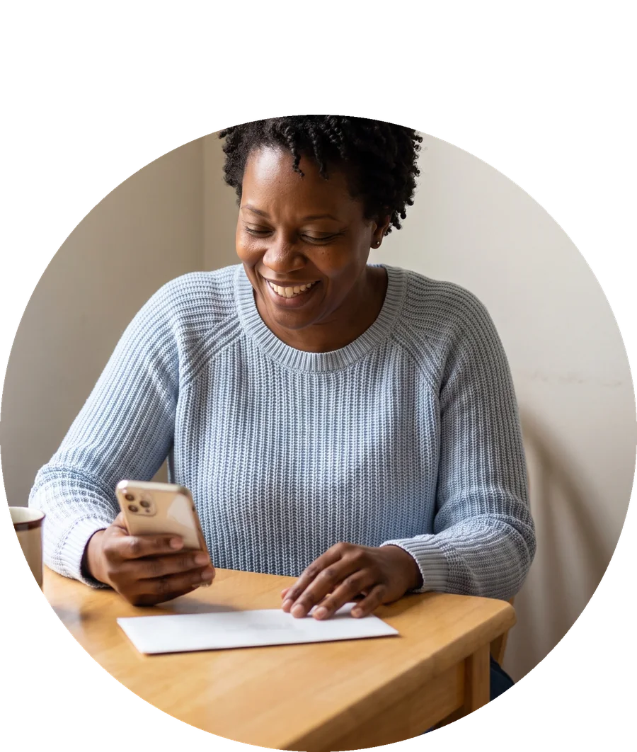 A smiling woman at her kitchen table checking her phone next to a bill